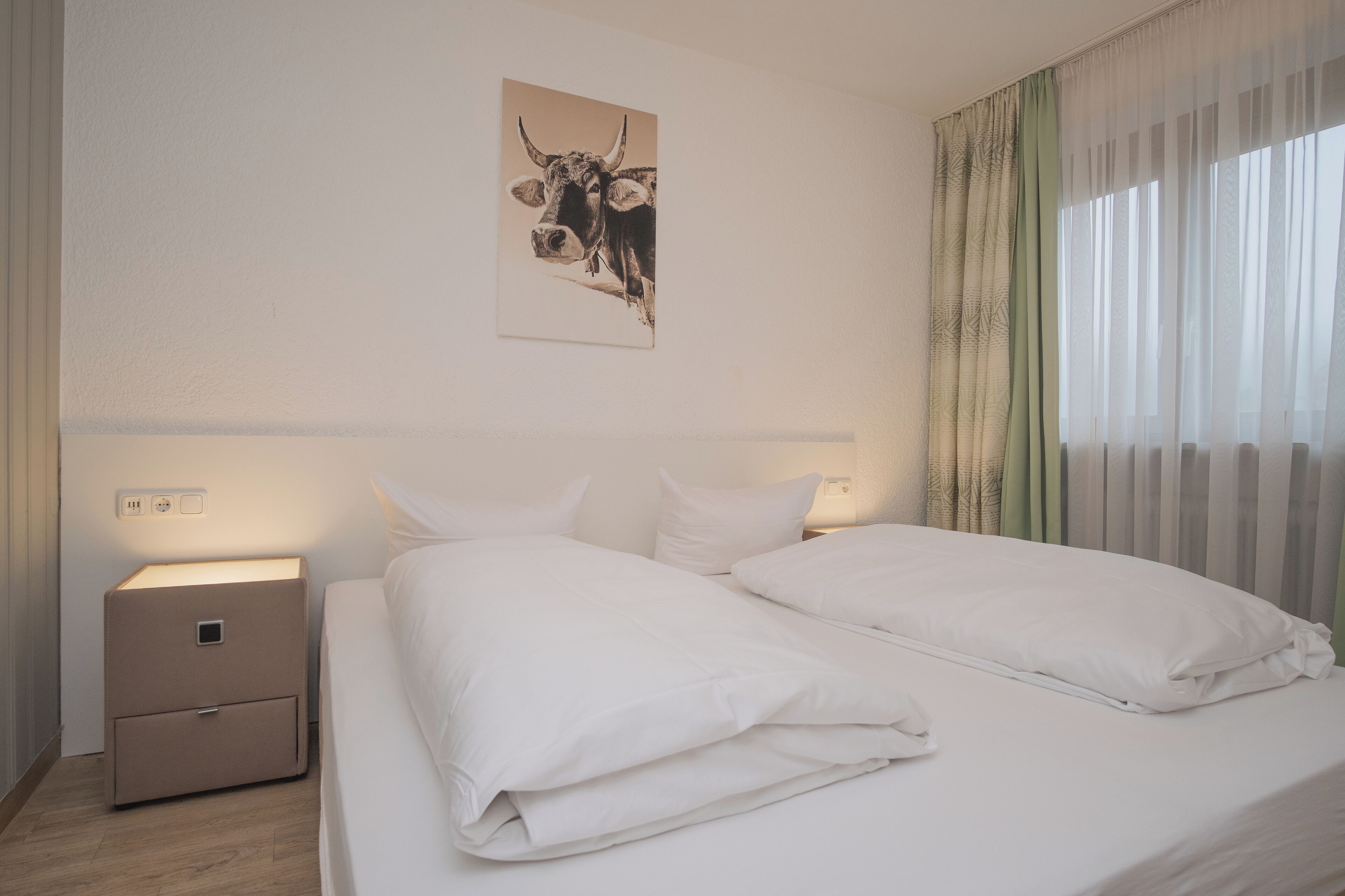 A hotel room with a bed, white bedding, a nightstand, a wall-mounted artwork of a bull, and sheer curtains.
