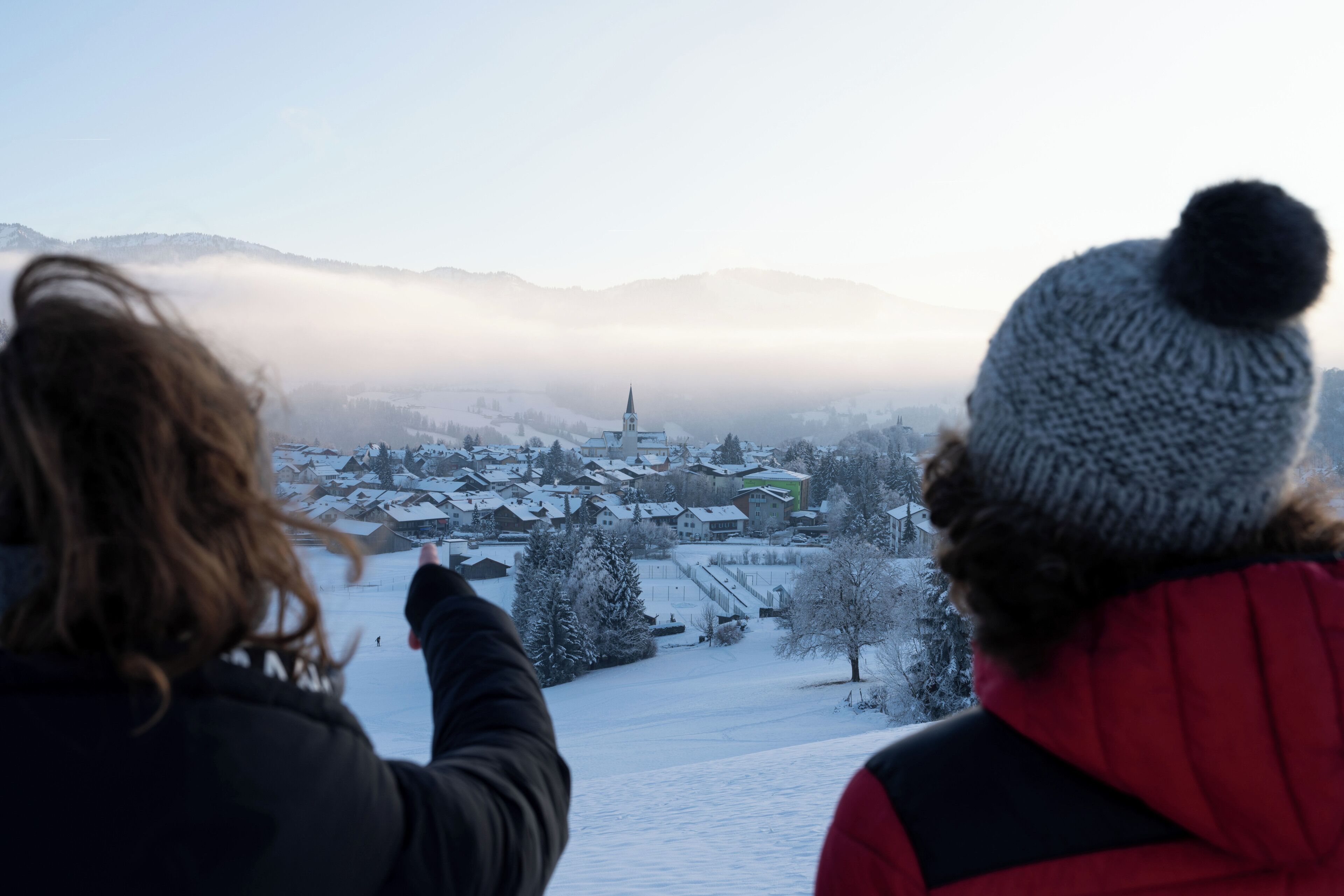 Two people overlooking a snowy village with a church spire, surrounded by mountains and fog.