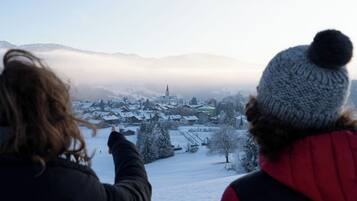 Zwei Personen blicken auf ein verschneites Dorf mit einem Kirchturm, umgeben von Bergen und Nebel.