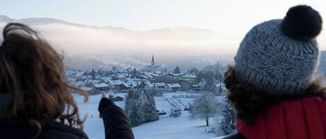 Two people overlooking a snowy village with a church spire, surrounded by mountains and fog.