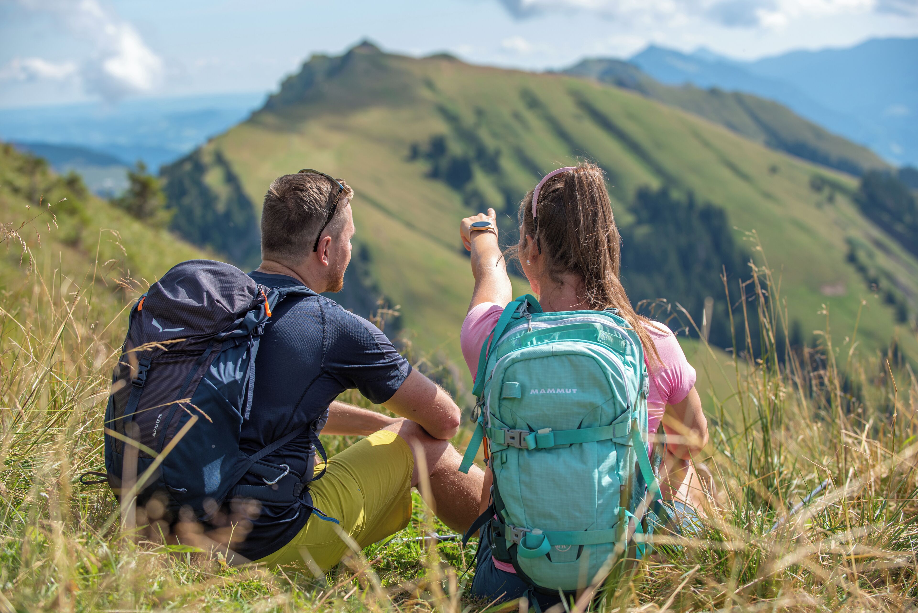 Two people sitting on grass with backpacks, overlooking a mountainous landscape.