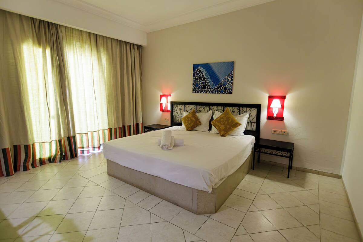 A hotel room with a large bed, two bedside tables, a headboard with a painting, and a window with curtains.