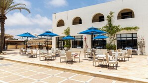 An outdoor seating area with tables and chairs, blue umbrellas, and a white building with arched windows. - Le Petit Palais Djerba & Spa (Djerba Midun)