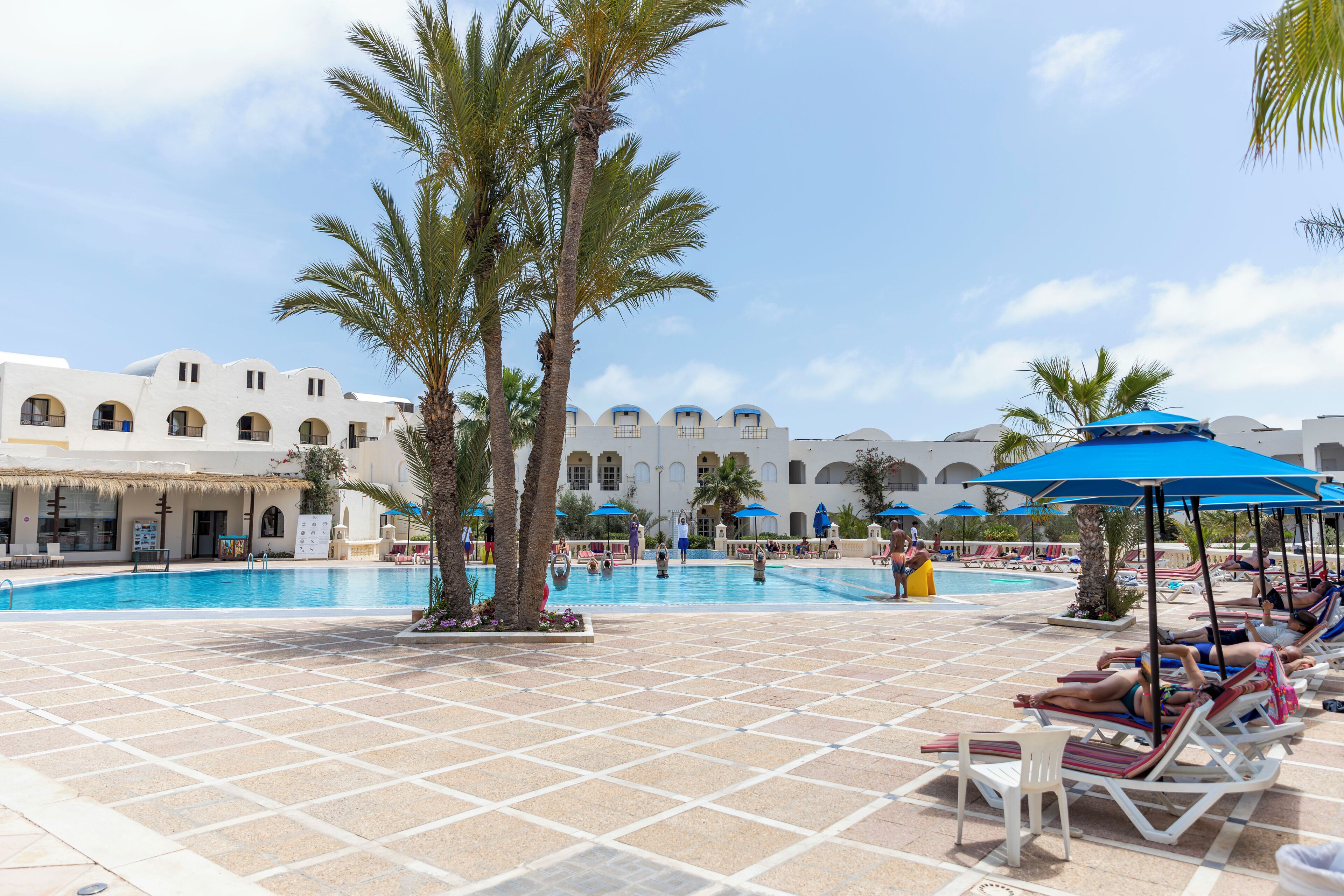A poolside area with lounge chairs and umbrellas, palm trees, and a building with arches in the background.