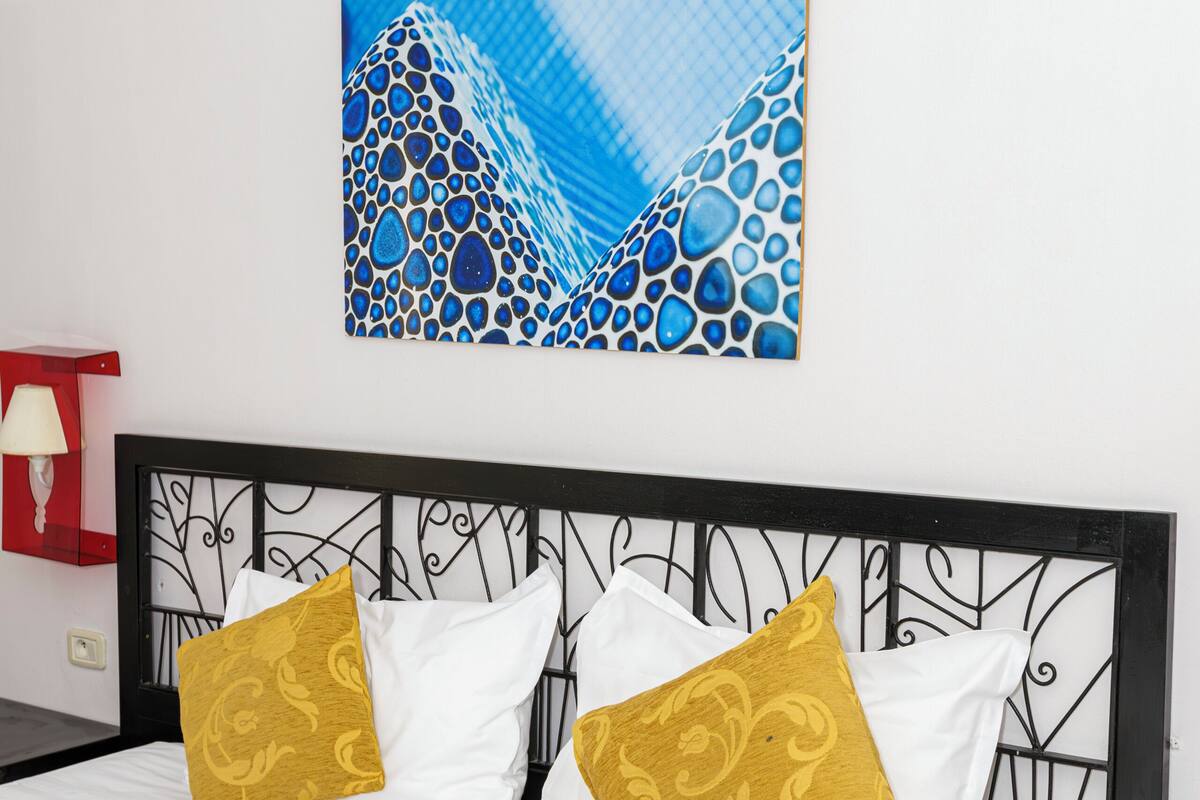A bed with white bedding and two gold-patterned pillows, a black metal headboard, and a blue abstract painting on the wall.