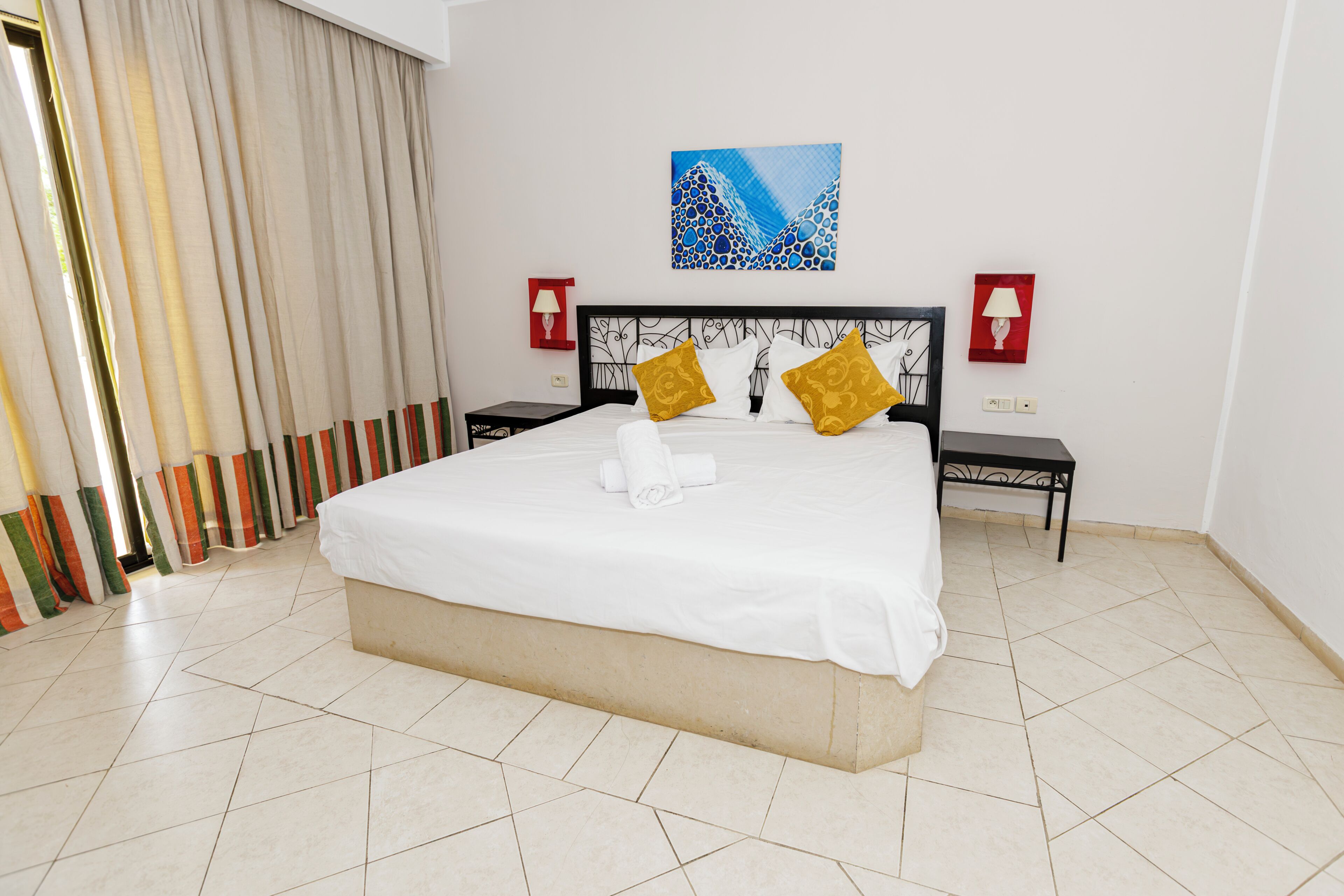 Un lit bien fait, avec du linge de lit blanc et deux coussins décoratifs, un tableau au mur et deux tables de chevet avec des lampes.