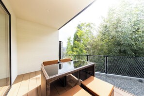 A balcony with a glass-top table and wicker chairs, overlooking a garden with trees and a fence. - TERRA Yufuin (Yufu)