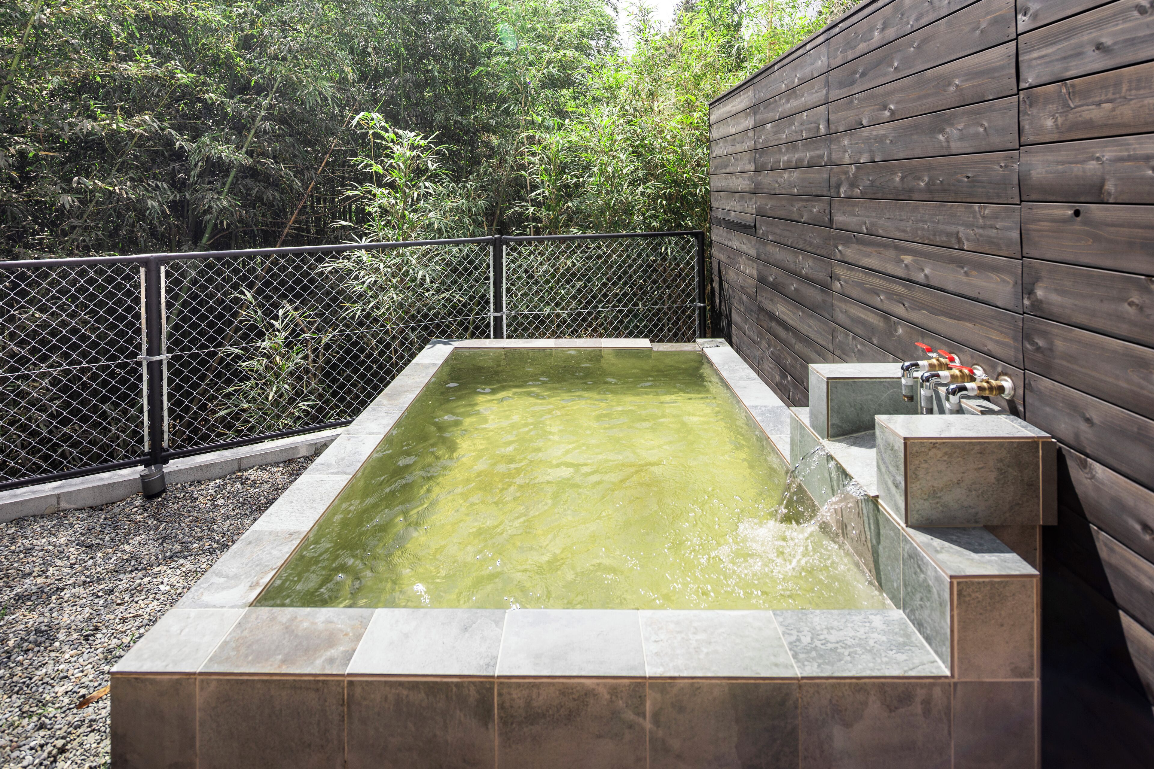 A rectangular hot tub with a wooden fence and a metal gate, surrounded by gravel and greenery.