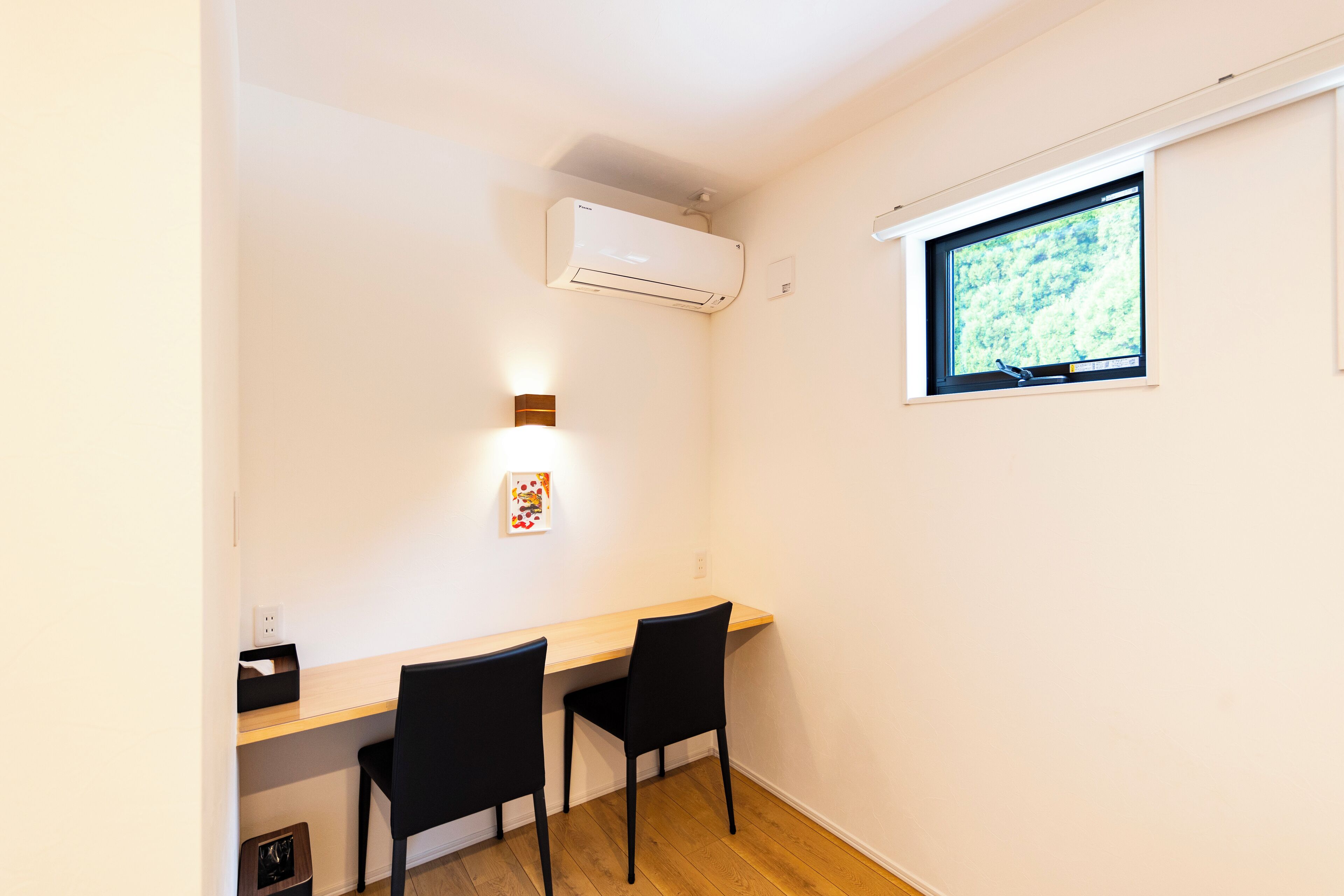 A small room with a wooden desk, two black chairs, a white air conditioner, a window with a view of greenery, and a wall-mounted light fixture.