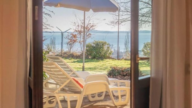A view from a room to an outdoor seating area with a table and chairs, a large umbrella, and a scenic lake view. - Modern and spacious for an unforgettable stay
 (Lazio)