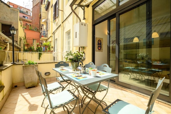 Eine Terrasse im Freien mit einem Tisch für zwei Personen, umgeben von Stühlen, einem Blumentopf und einem großen Fenster mit Blick auf die Straße.