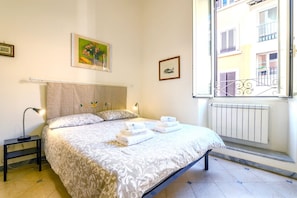 Ein Schlafzimmer mit einem Bett, einem Nachttisch, einem Fenster mit Blick auf Gebäude und einer Heizung.