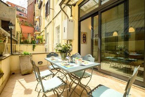 An outdoor patio with a table set for two, surrounded by chairs, a potted plant, and a large window with a view of the street.