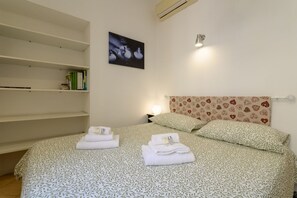 Een netjes opgemaakt bed met handdoeken, een boekenkast, een aan de muur bevestigd schilderij en een bed met een decoratief hoofdeinde.