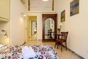 A hotel room with a bed, a wooden chair, a table with a plant, a mirror, and a painting on the wall.