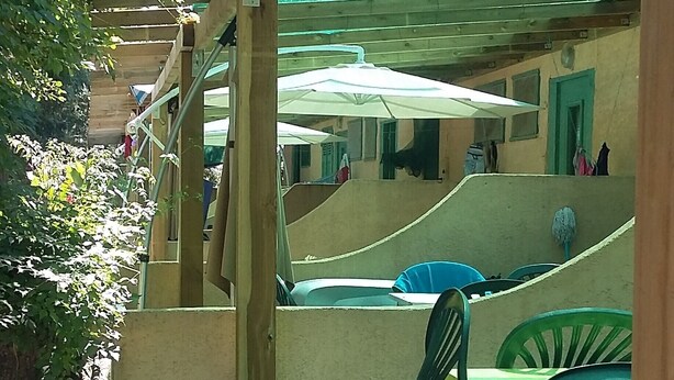 Ett takförsett utomhusområde med sittplatser, en träpergola, ett betonggolv och gröna plaststolar.