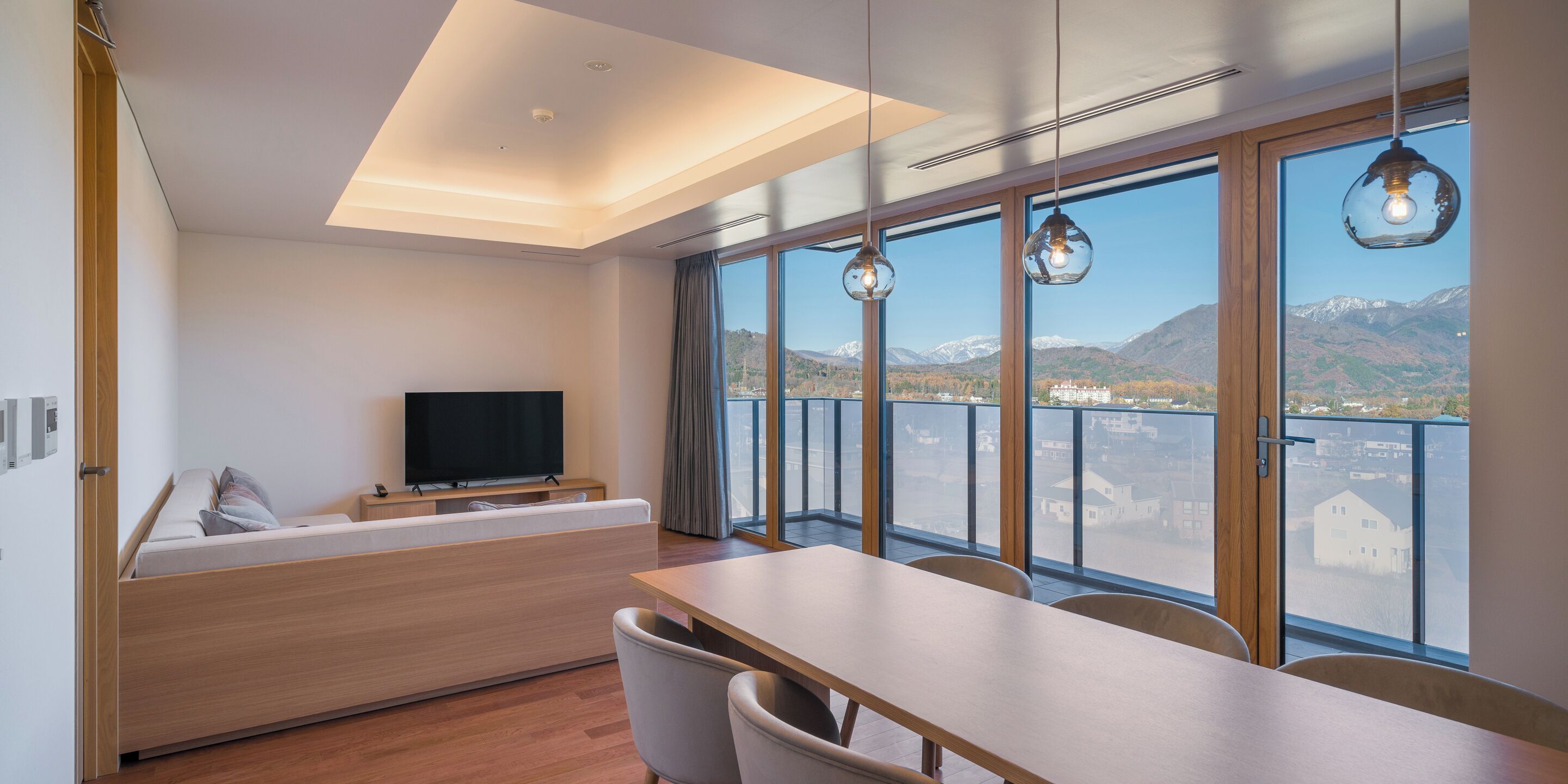 Una habitación de hotel moderna con una cama de madera, un televisor, una mesa de comedor y ventanas grandes con vistas a las montañas.