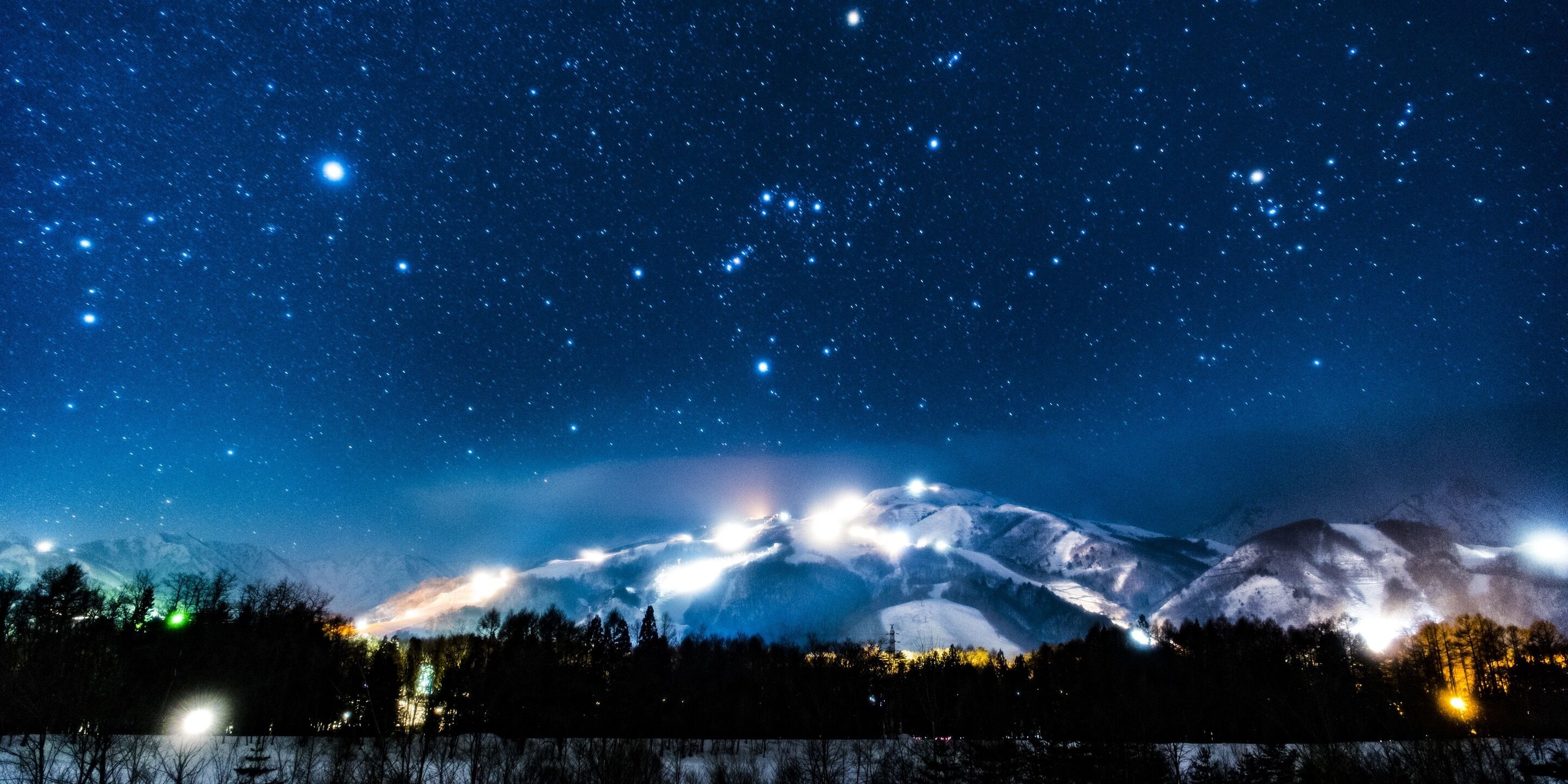 A mountain range under a starry night sky with snow-covered peaks and a forested foreground.