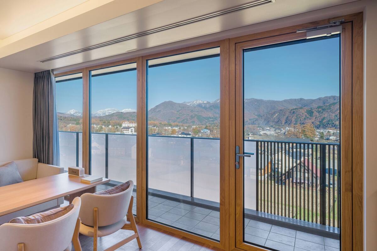 A modern living room with a sofa, a dining table, and chairs. It features large windows with a mountain view.