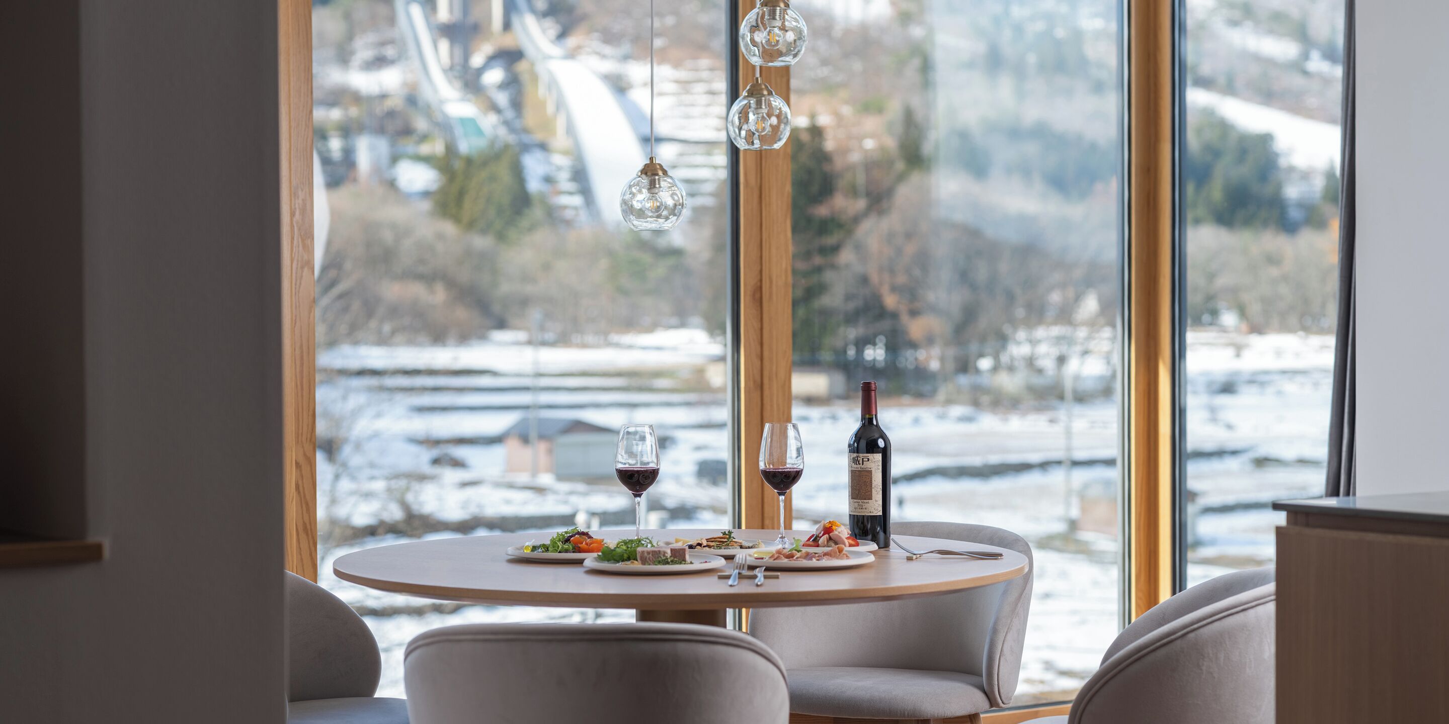 Una mesa de comedor con platos de comida, copas de vino y una botella de vino, con vistas a un paisaje nevado a través de una ventana.