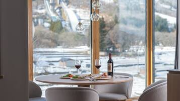 Una mesa de comedor con platos de comida, copas de vino y una botella de vino, con vistas a un paisaje nevado a través de una ventana.