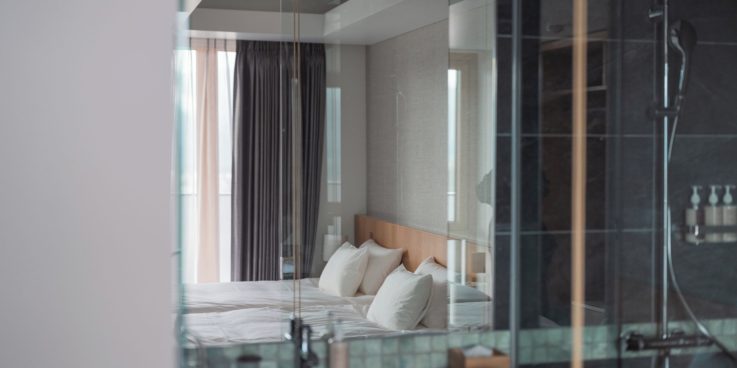 Quarto de hotel moderno com uma cama grande, chuveiro em cabine de vidro e vista para o exterior pela janela.