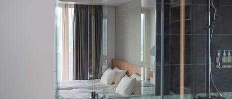 Quarto de hotel moderno com uma cama grande, chuveiro em cabine de vidro e vista para o exterior pela janela.