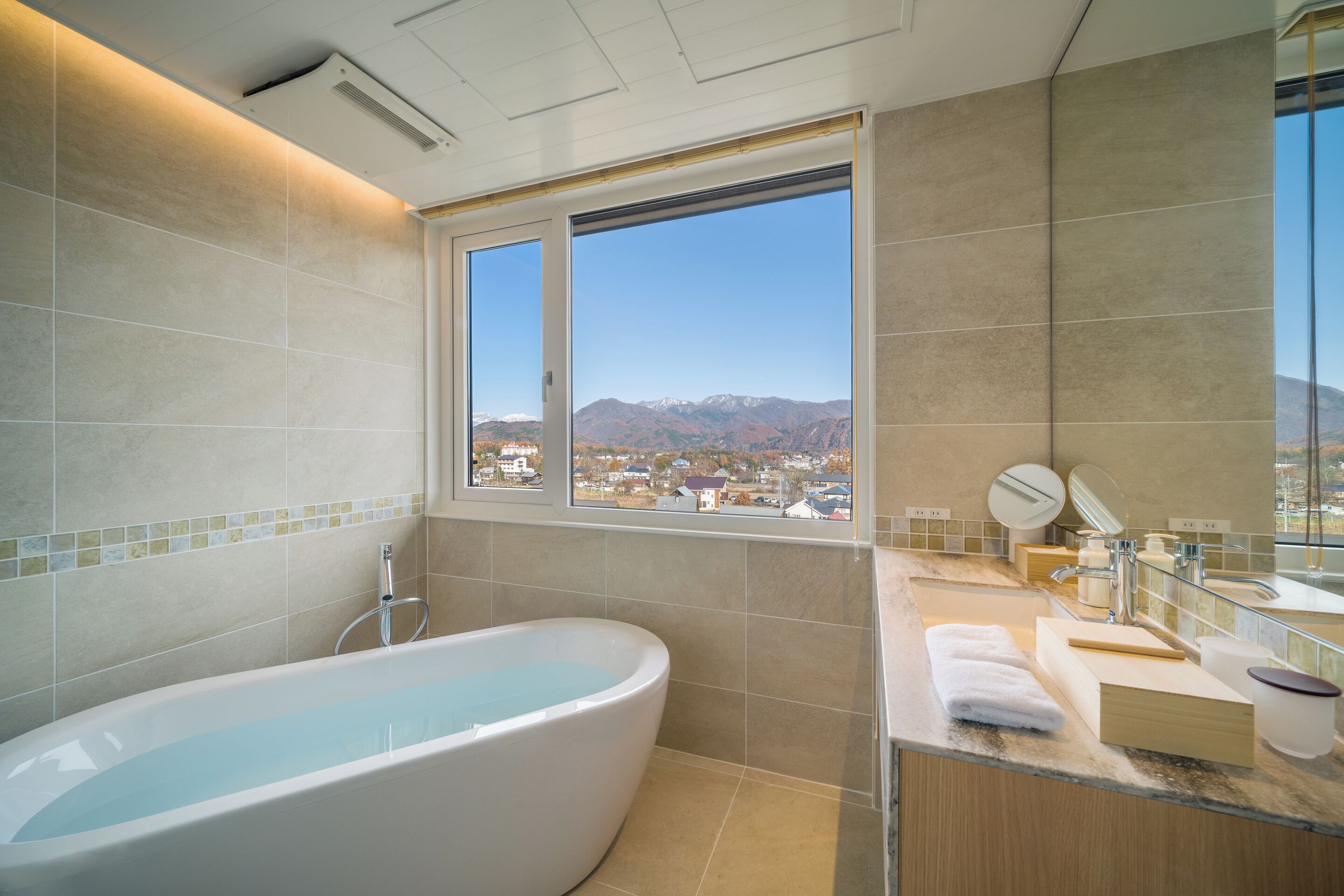 Une salle de bain moderne avec une grande baignoire, un meuble-lavabo avec un miroir et une vue sur la ville par la fenêtre.
