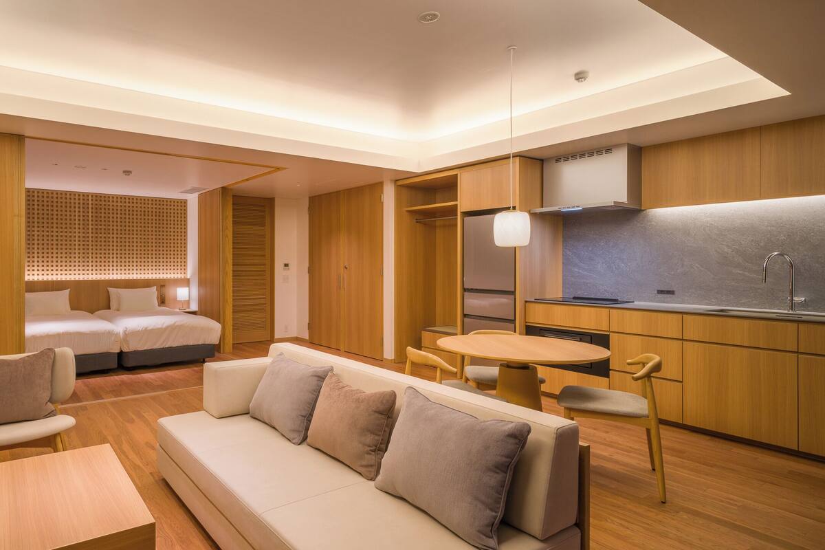 A modern hotel room with a sofa, a dining table, a kitchenette, and a separate sleeping area.