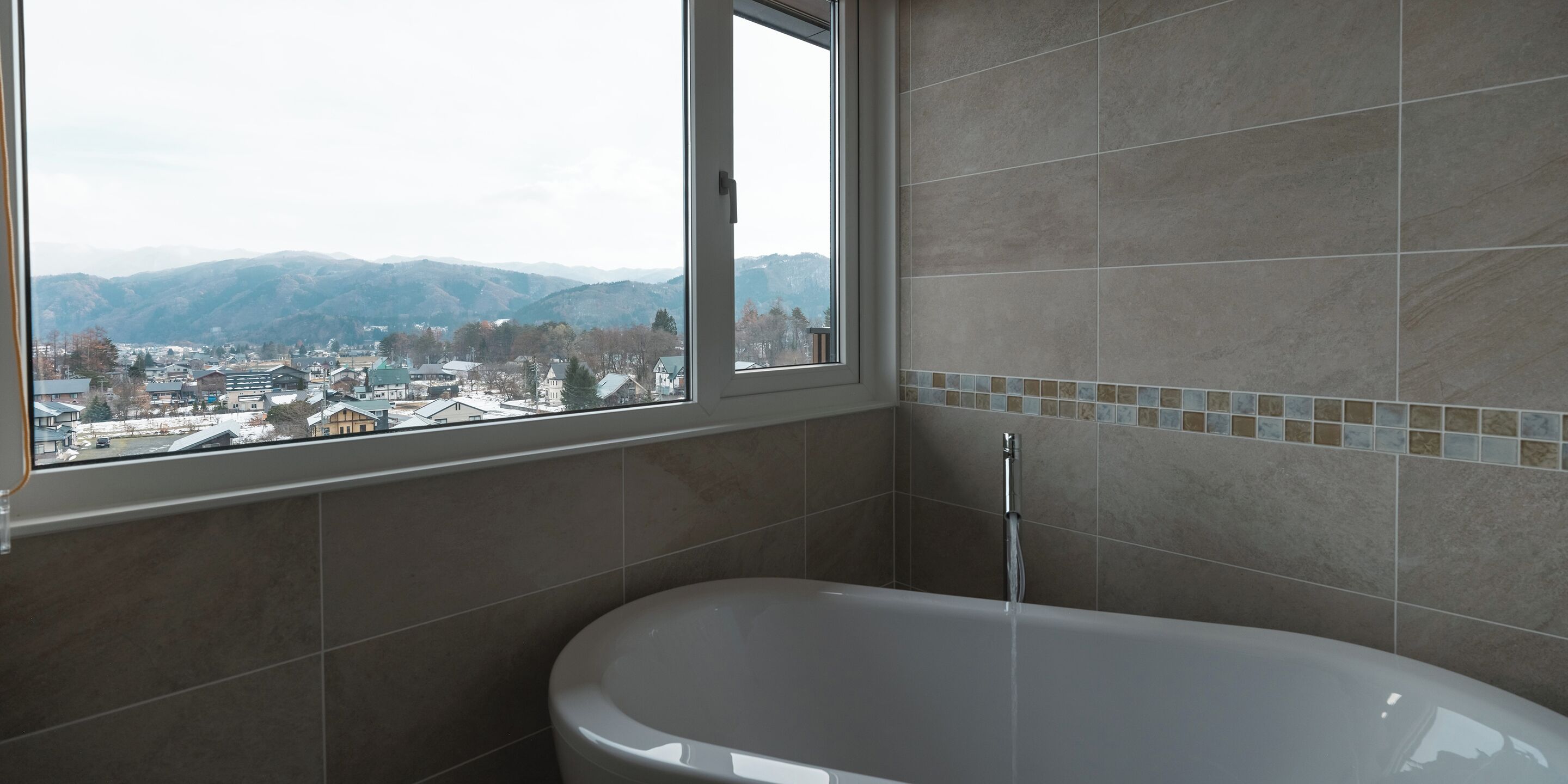 A bathroom with a bathtub, a window with a view of a town and mountains, and tiled walls.