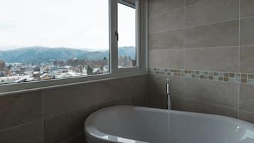 A bathroom with a bathtub, a window with a view of a town and mountains, and tiled walls.