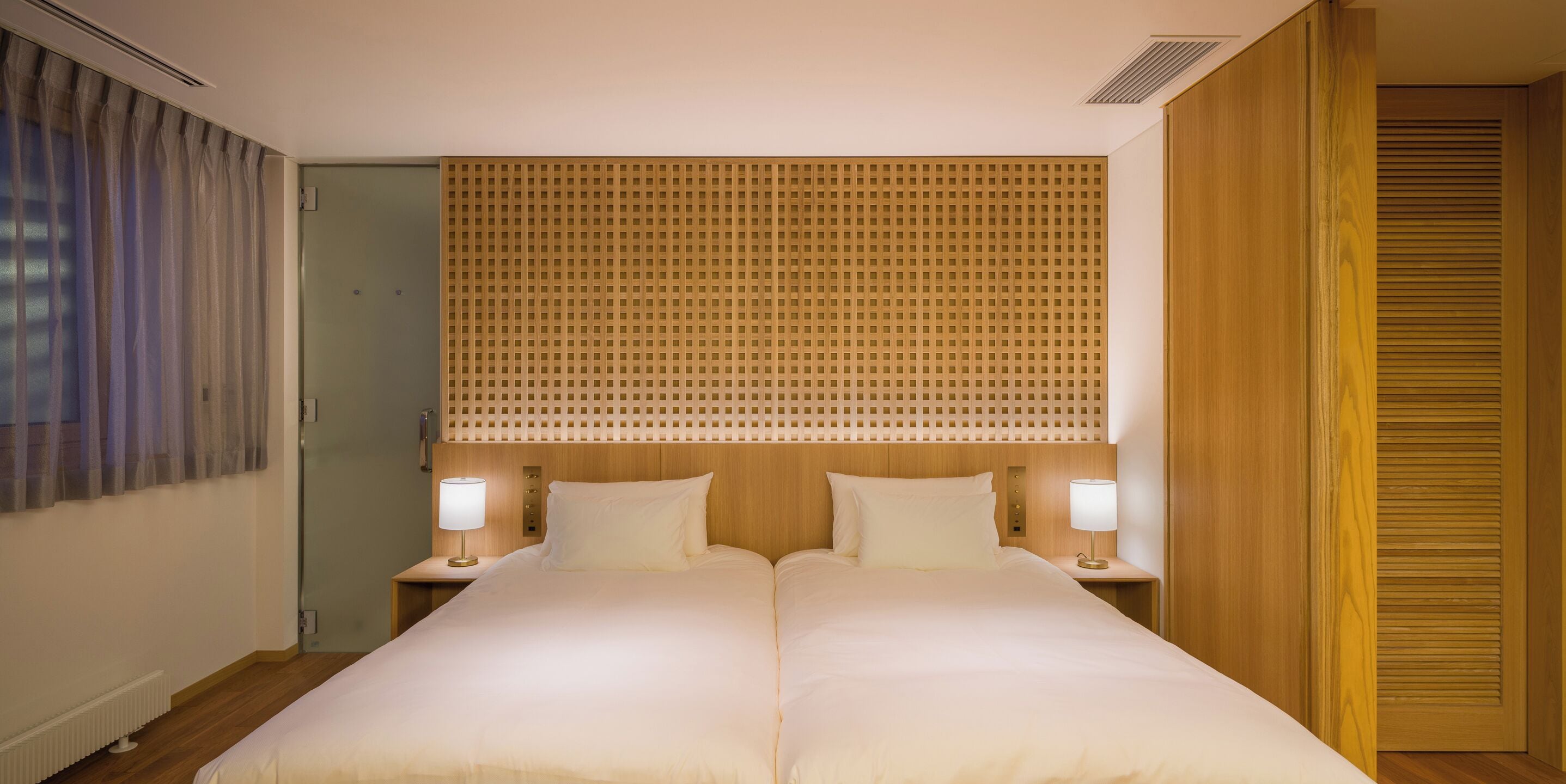 A double bed with two nightstands, each with a lamp, in a room with wooden paneling and a door to the left.