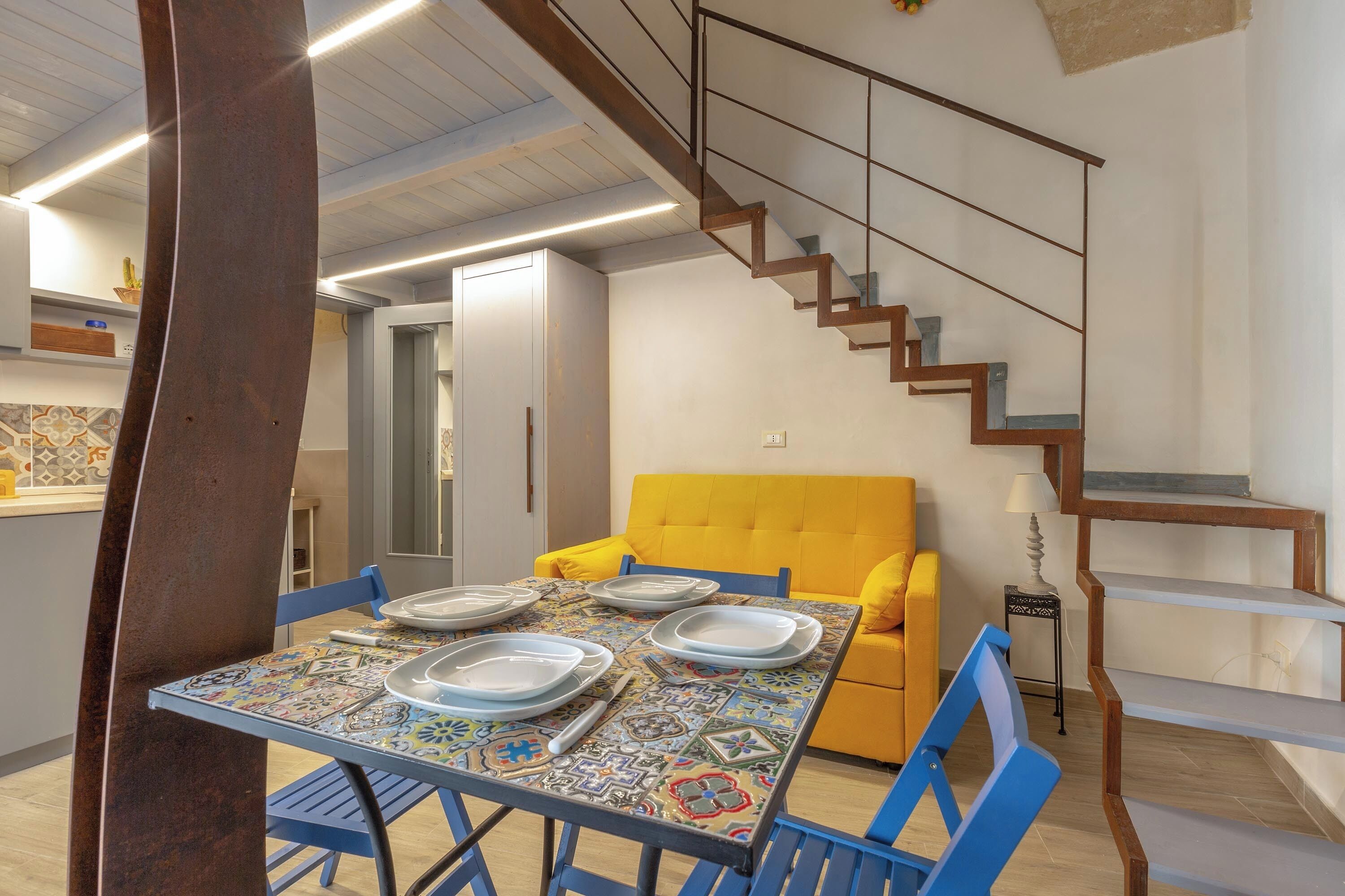 Un comedor con una mesa preparada para cuatro personas, un sofá amarillo y una escalera.