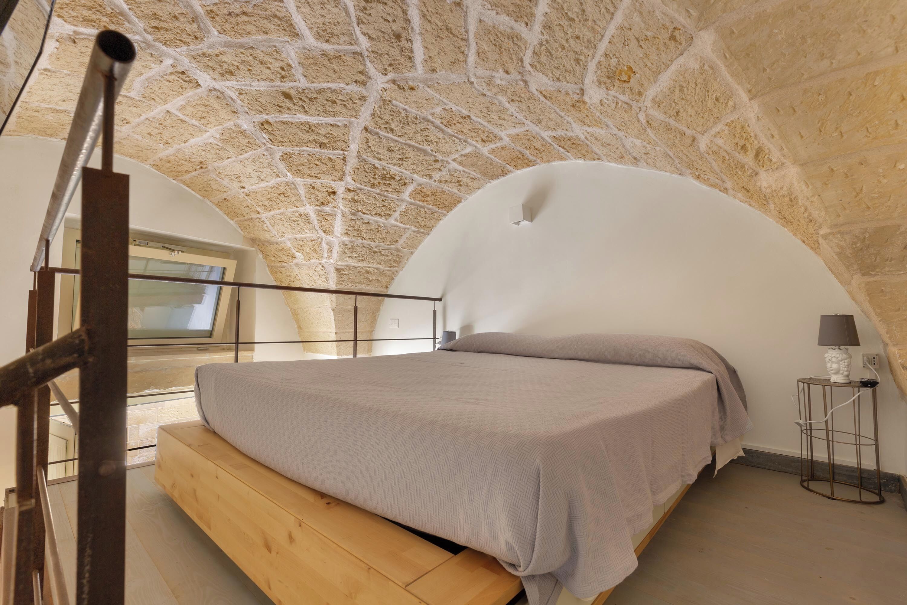 A bedroom with a stone ceiling, a bed with a wooden platform, and a small table with a lamp.