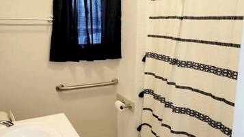 Un baño con lavamanos blanco, cortina de ducha a rayas negras y blancas, y una ventana con cortinas negras.