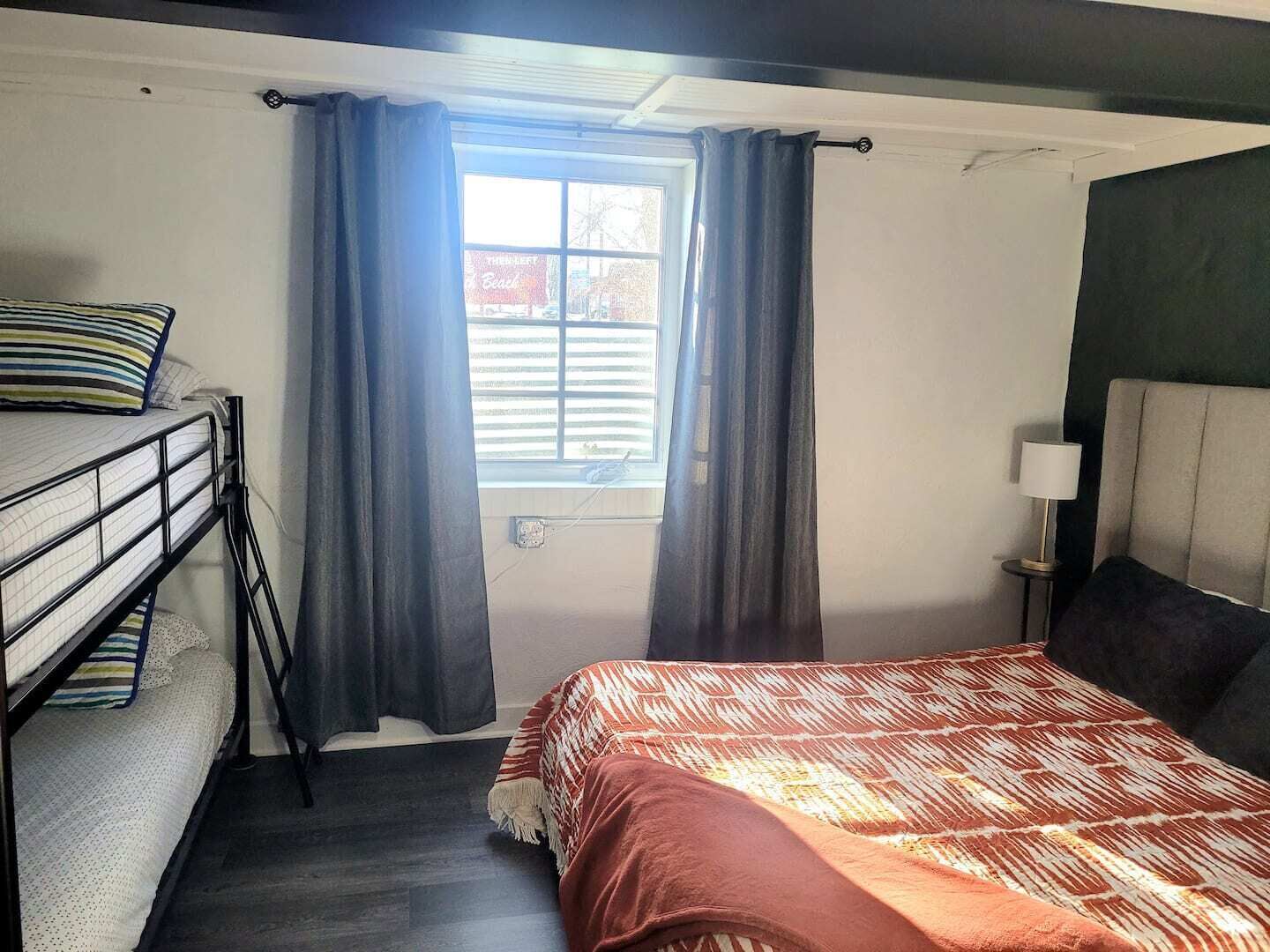 A room with a bunk bed, a single bed, a window with curtains, a bedside lamp, and a view of the outside.