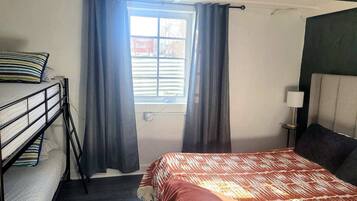 A room with a bunk bed, a single bed, a window with curtains, a bedside lamp, and a view of the outside.