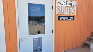 A hotel entrance with a sign reading "stay SUITES" and a website address "STAYBOJI.COM". There is a door with a window showing a beach view, and a sign indicating directions to various locations.