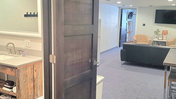 A modern hotel room with a large dark wooden door, a bathroom with a sink and mirror, a living area with a sofa and TV, and a carpeted floor.
