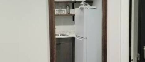 Un couloir étroit, orné d’un grand miroir au mur, mène à une cuisine équipée d’un réfrigérateur blanc et de placards.