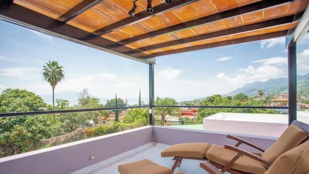 Un balcón con vistas a zonas verdes y edificios, equipado con sillas de madera y una pérgola.