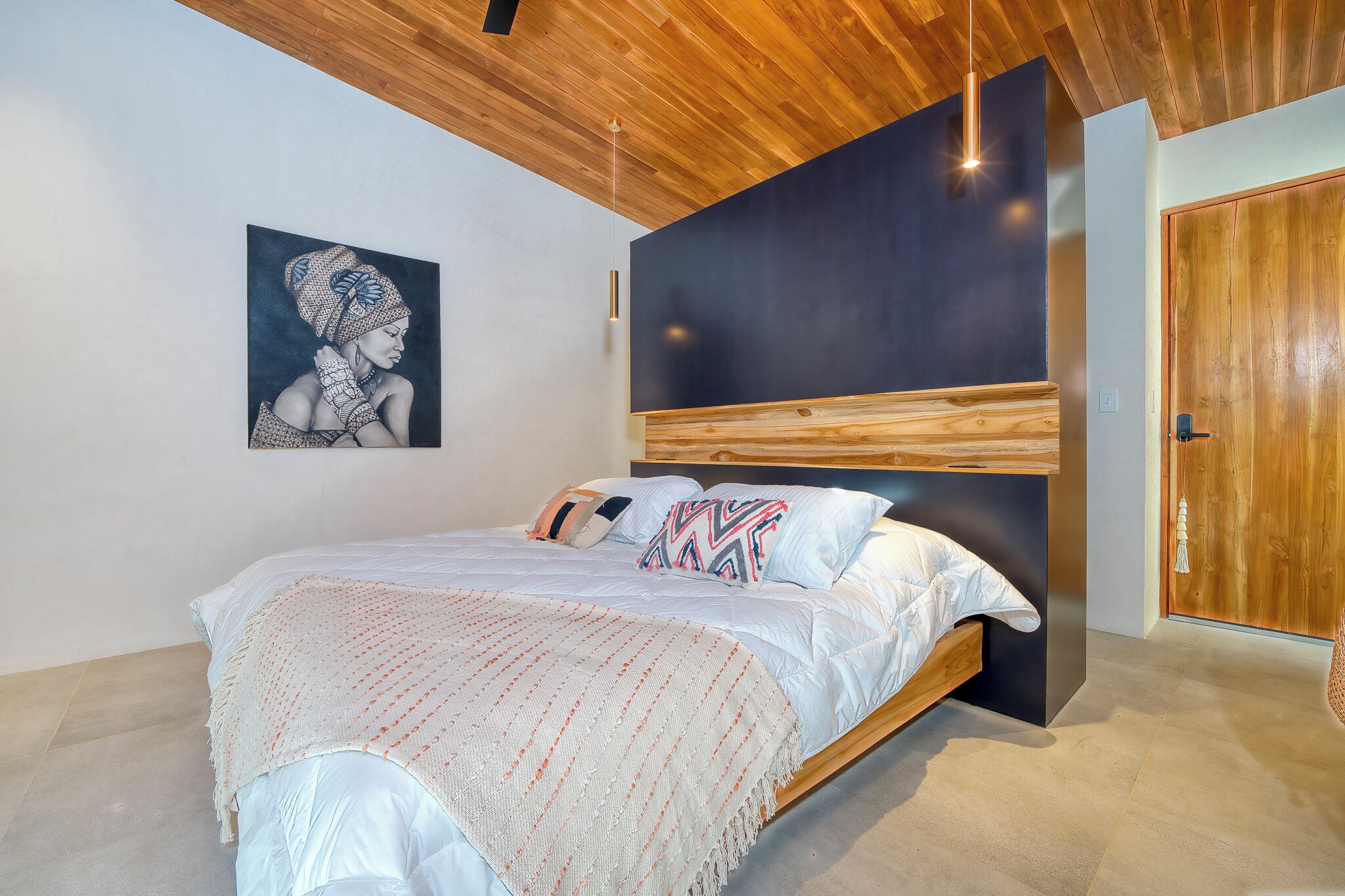 Un dormitorio moderno con cabecera de madera, una cama grande con ropa blanca y almohadas estampadas, una obra de arte colgada en la pared y una puerta de madera.