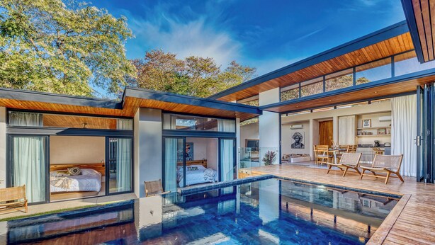 A modern house with a glass-enclosed bedroom, a pool, and a dining area with wicker furniture.
