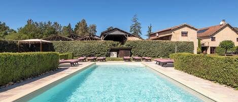 A rectangular swimming pool with lounge chairs and a covered area on one side, surrounded by hedges and trees.