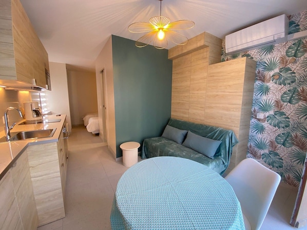 A compact hotel room with a kitchenette, a round table, a sofa, and a patterned wall.