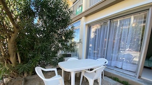 A patio area with a white table and chairs, surrounded by greenery and a building with a large window. - Cozy apartment with private garden near thermal baths (Balaruc-les-Bains)