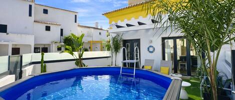 Una piscina con escalera, rodeada de edificios blancos con detalles amarillos, y una pequeña área ajardinada con plantas.