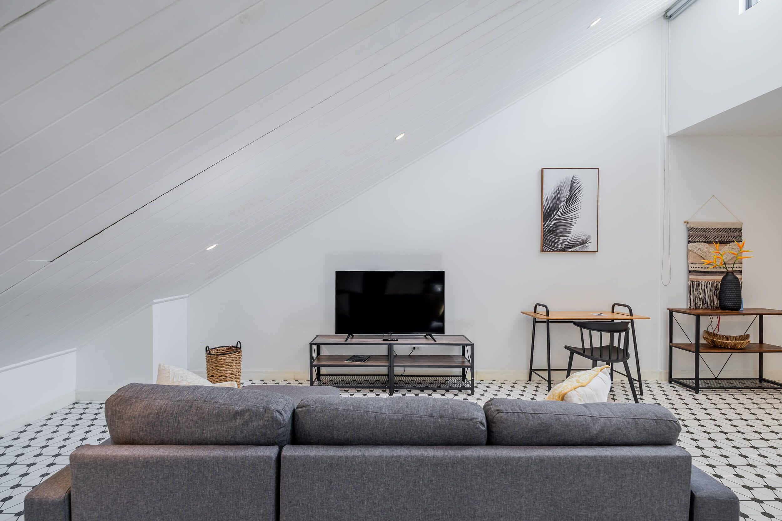 En moderne stue i loftstil med et skakternet gulv i sort og hvid, en grå sofa, et fjernsyn på et træstel, et lille spisebord og en vægmonteret hylde med dekorative genstande.