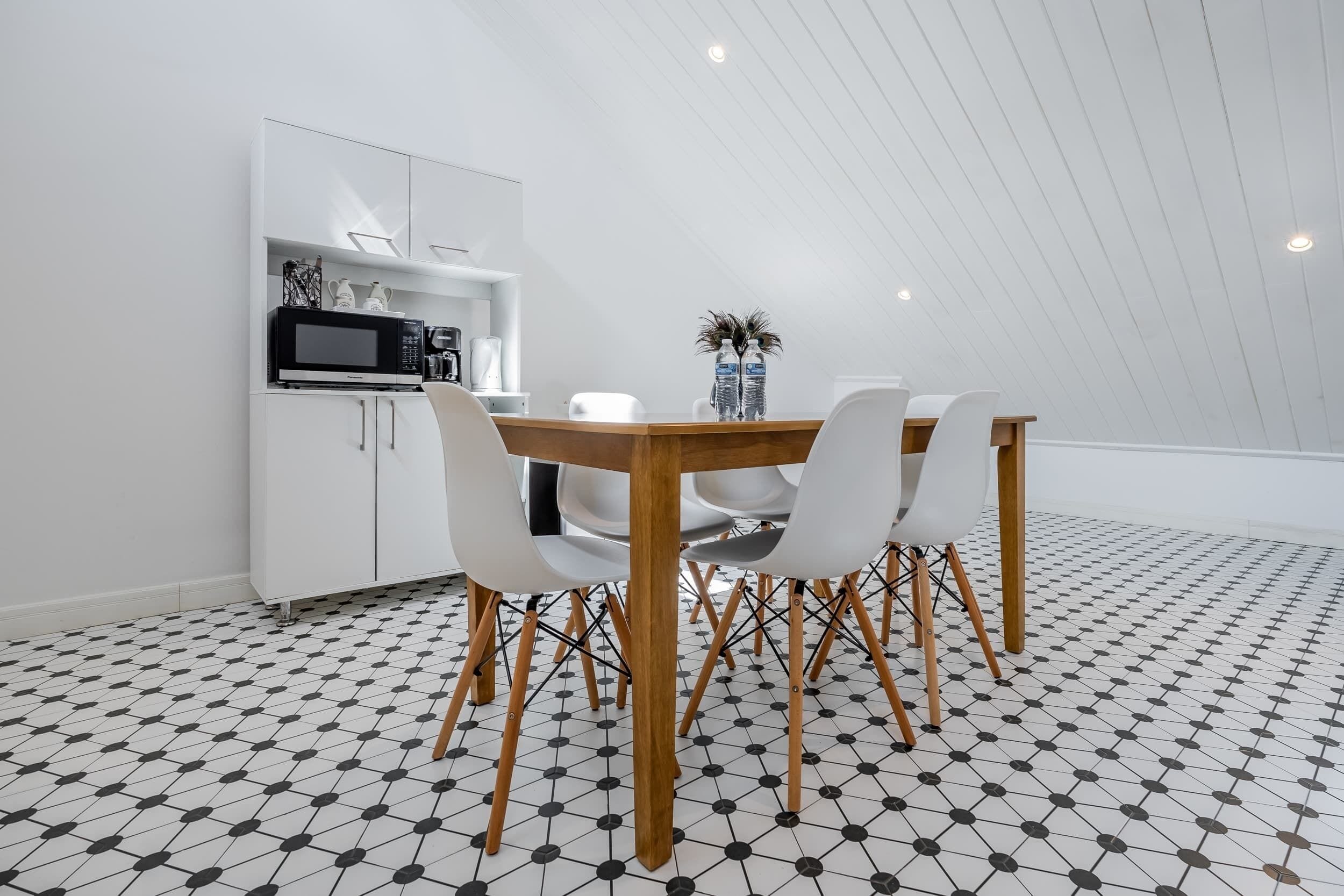 Een eethoek met een houten tafel en witte stoelen, een witte kast met een magnetron, en een betegelde vloer.