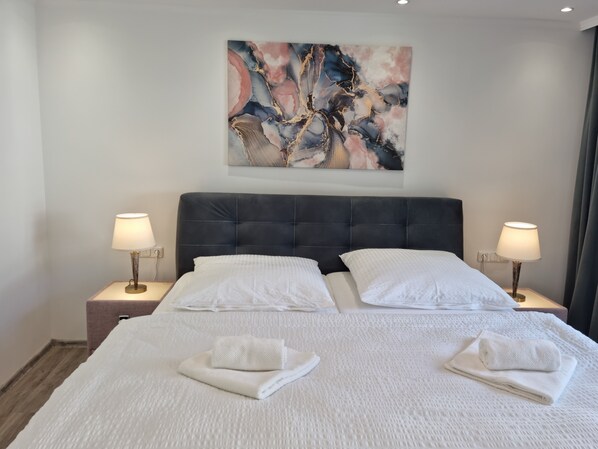 Ein Doppelbett mit weißen Leinentüchern und zwei gefalteten Handtüchern, mit Nachttischen und Lampen an beiden Seiten, in einem Zimmer mit einem großen, abstrakten Gemälde an der Wand.